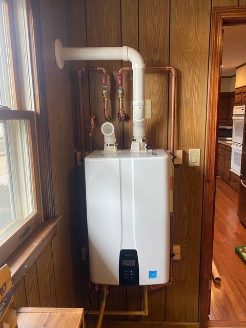 White tankless water heater mounted on wooden wall with copper and PVC pipes, digital control panel, and window visible to the left