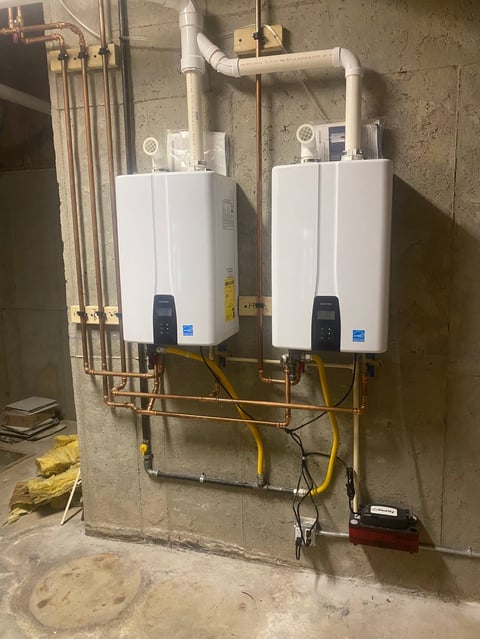 Two white tankless water heaters mounted on a concrete basement wall with copper and PVC piping connections above