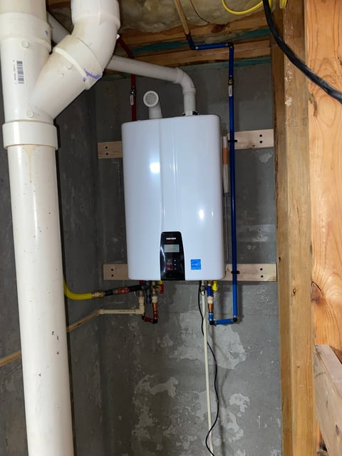 Tankless water heater mounted on concrete wall with white and blue PVC piping and electrical connections in a basement utility room