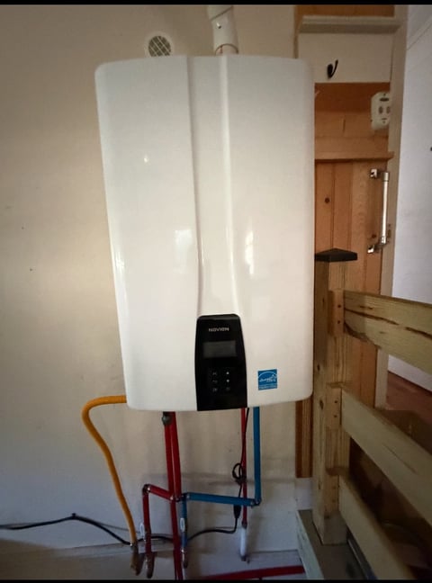 White tankless water heater mounted on wall with black control panel and colored supply pipes in a utility room