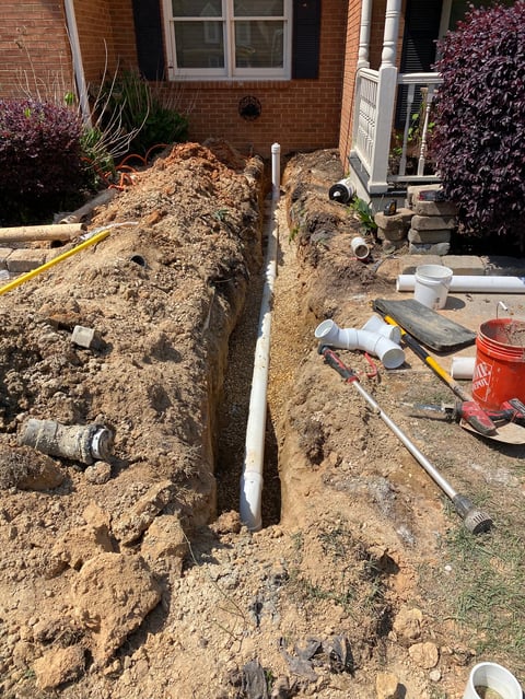 Excavated trench next to brick house with underground pipes and plumbing materials during construction or repair work