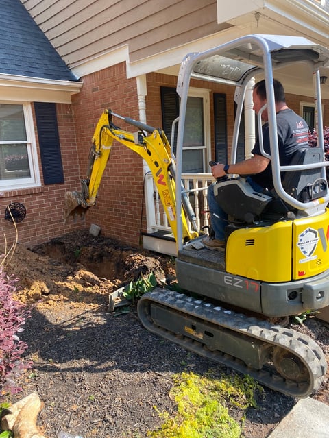 Operator in yellow and gray compact excavator excavating soil near brick house foundation during construction work